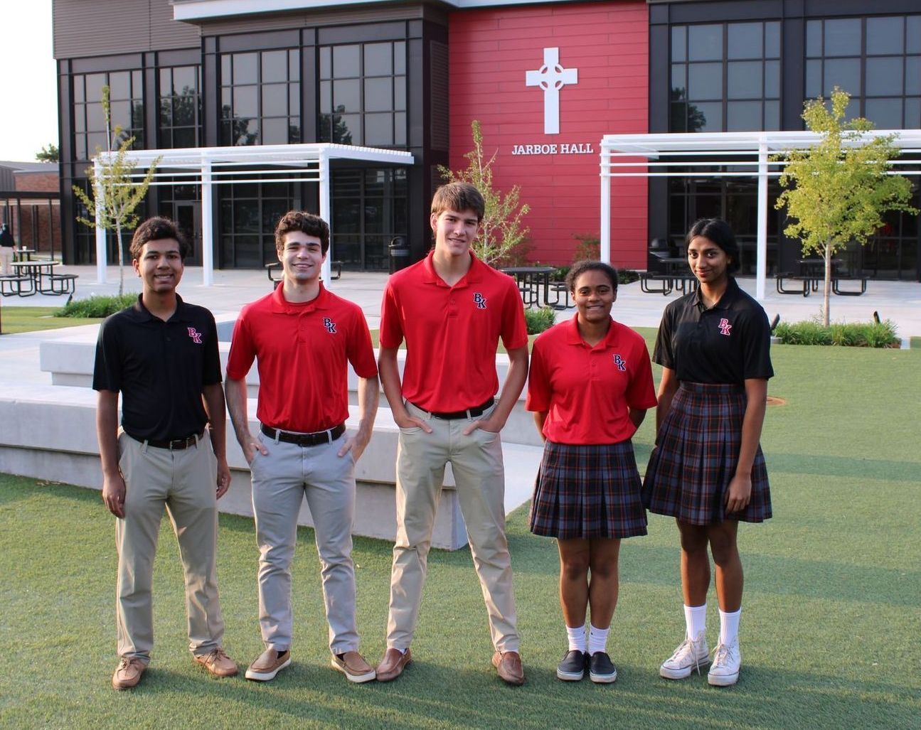 Five students in school uniforms stand outside a building with a cross symbol.