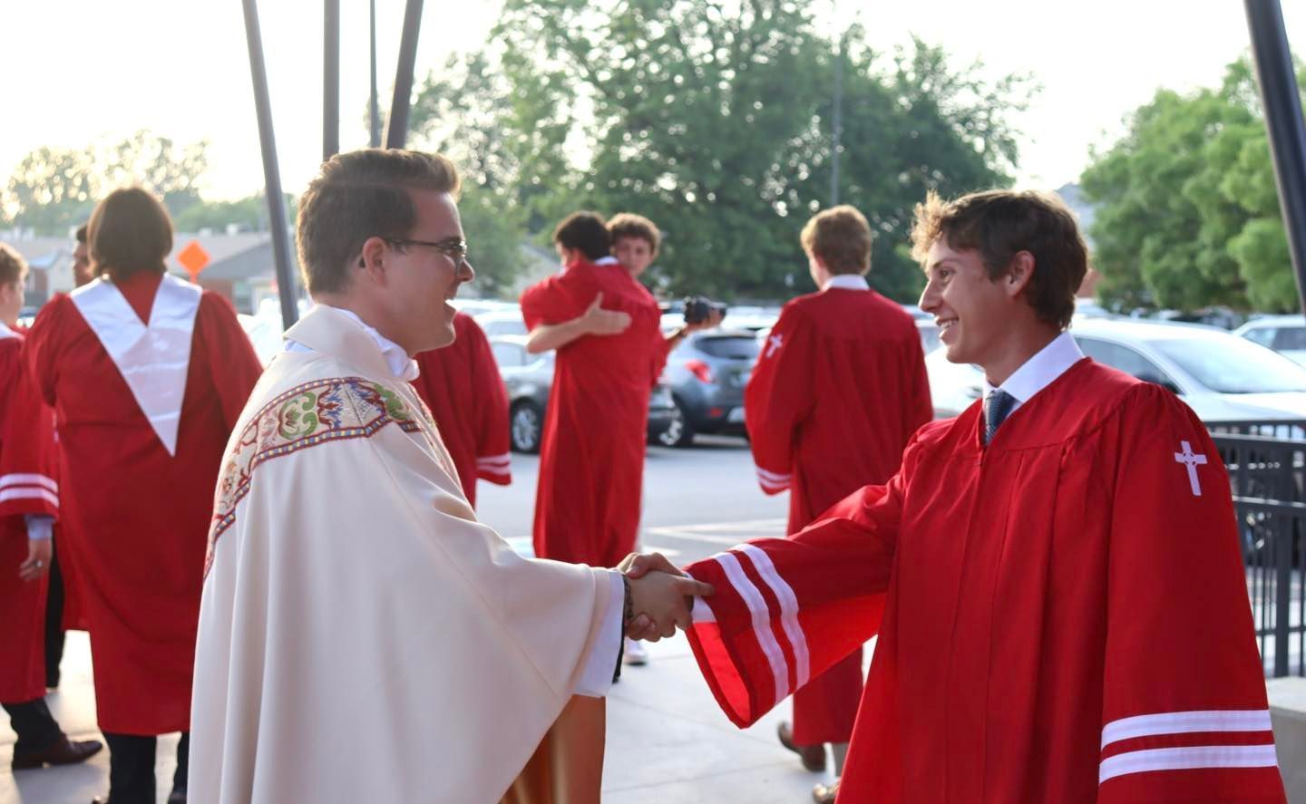 A priest shakes hands with a person in a red robe, others in robes, exterior of a building.