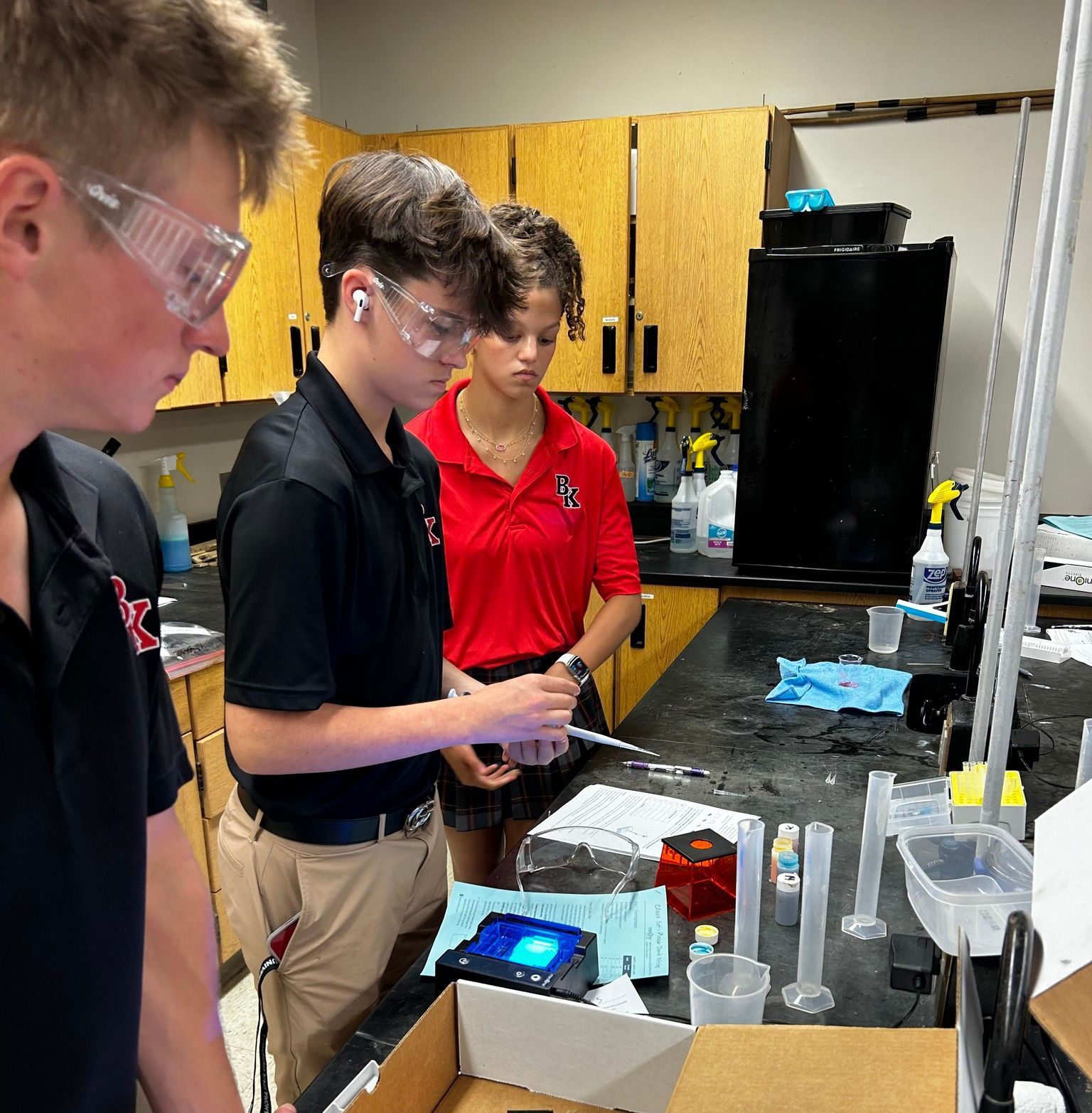 Three students wearing safety glasses conduct a science experiment in a lab.