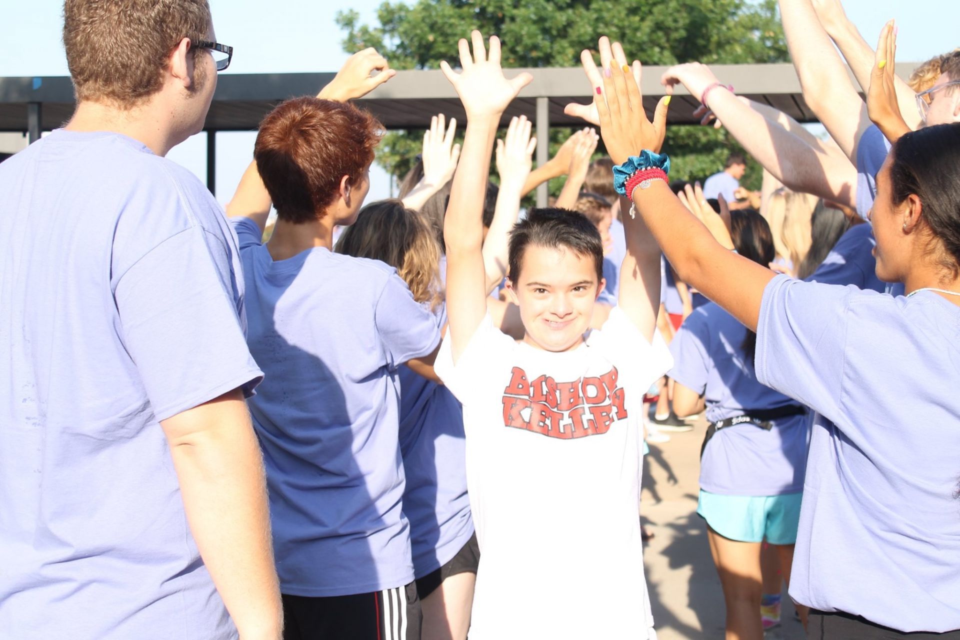 Children in blue shirts high-five a smiling boy outdoors in bright sunlight