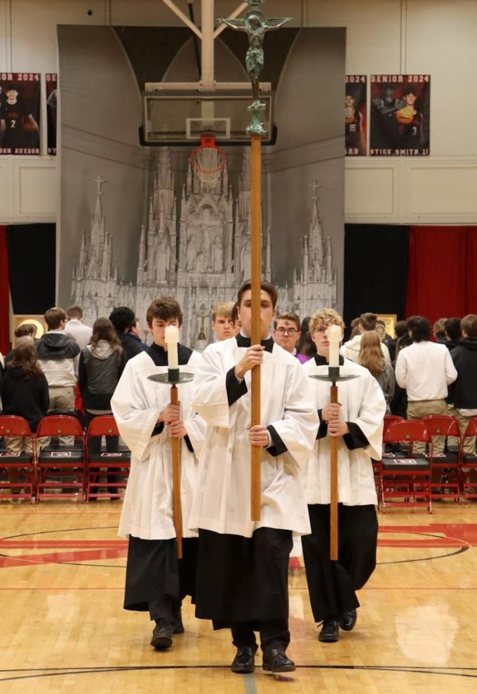Altar servers lead a procession carrying candles and a crucifix in a gymnasium. People in the background.