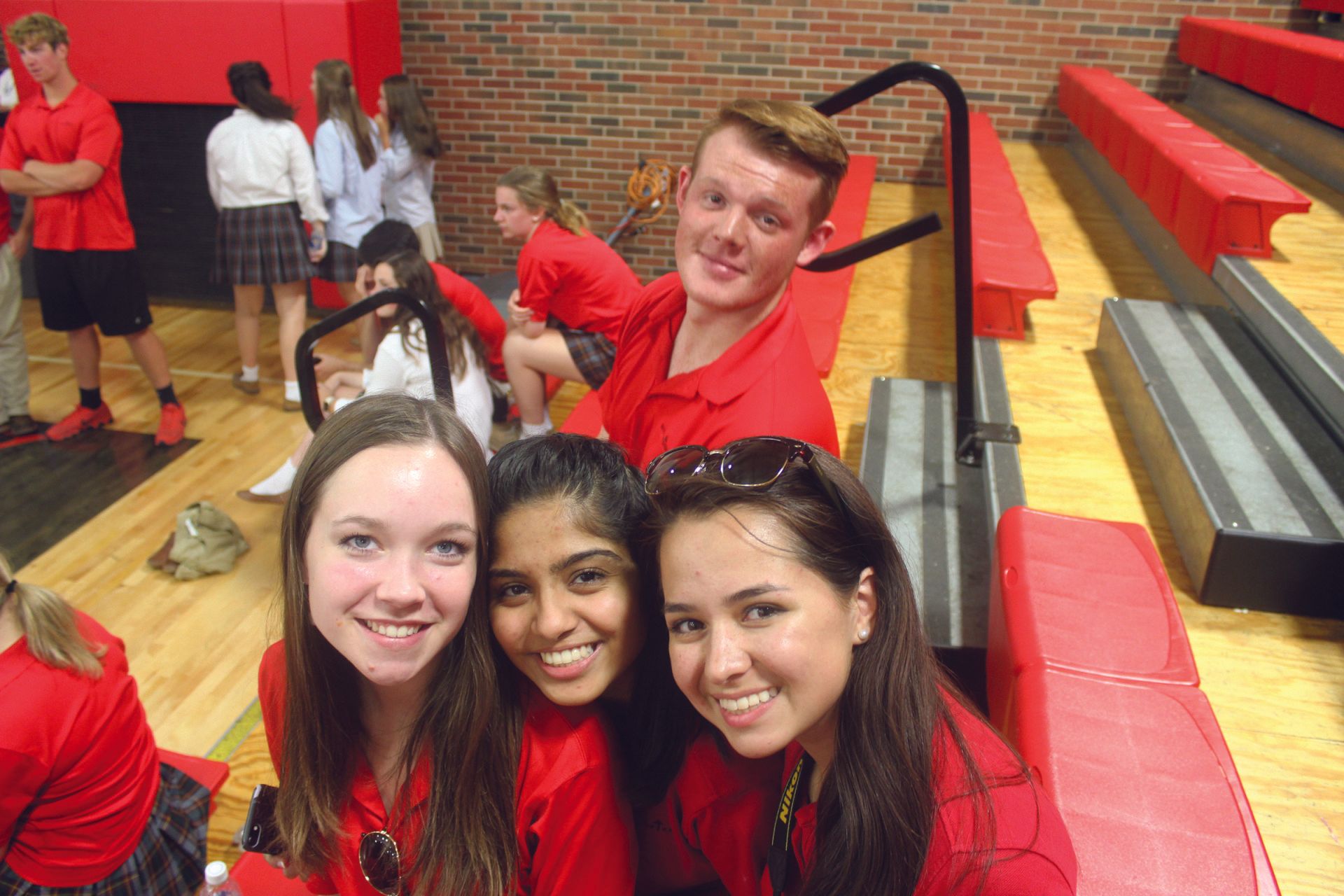 Four smiling students in red shirts taking a selfie in a gym with red bleachers