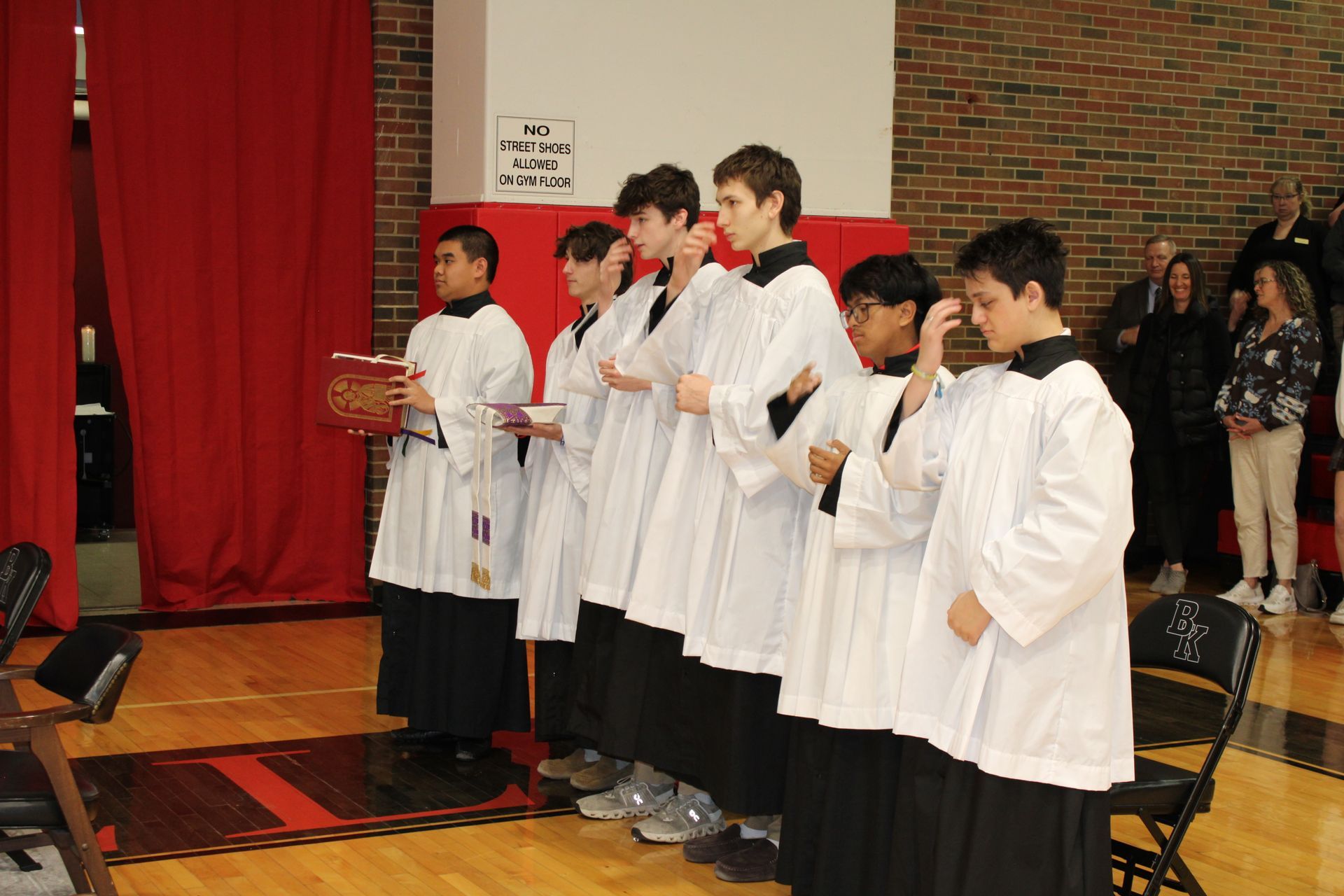 Priest holding chalice during service, surrounded by students in a school setting.