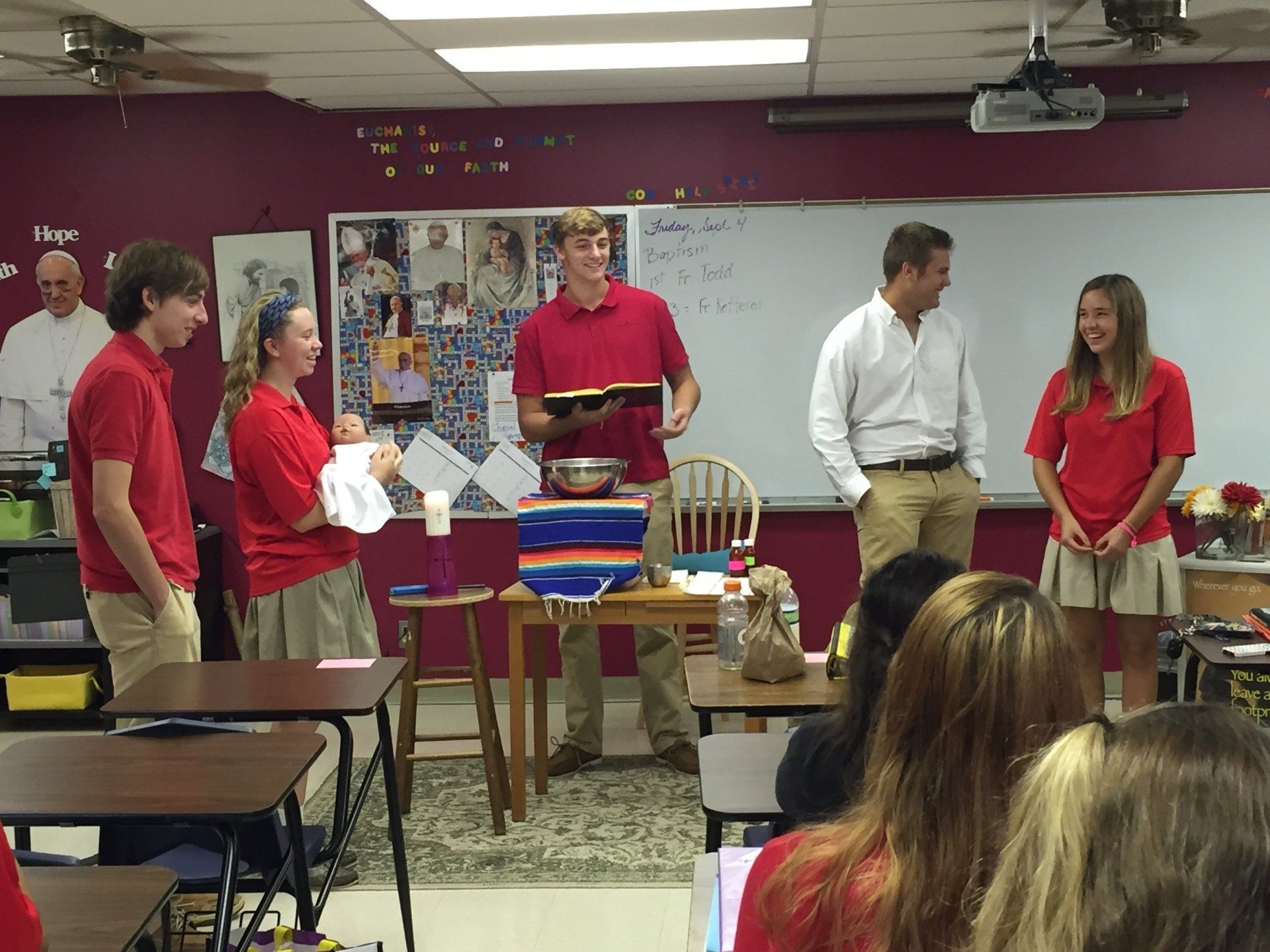 People standing in a classroom beside a table with gifts and a cake, as one person speaks.