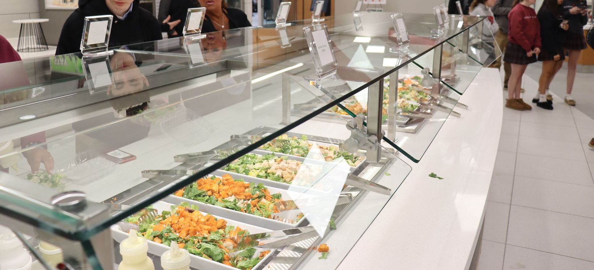A salad bar with various ingredients behind a glass display case. People are at the counter, selecting food.