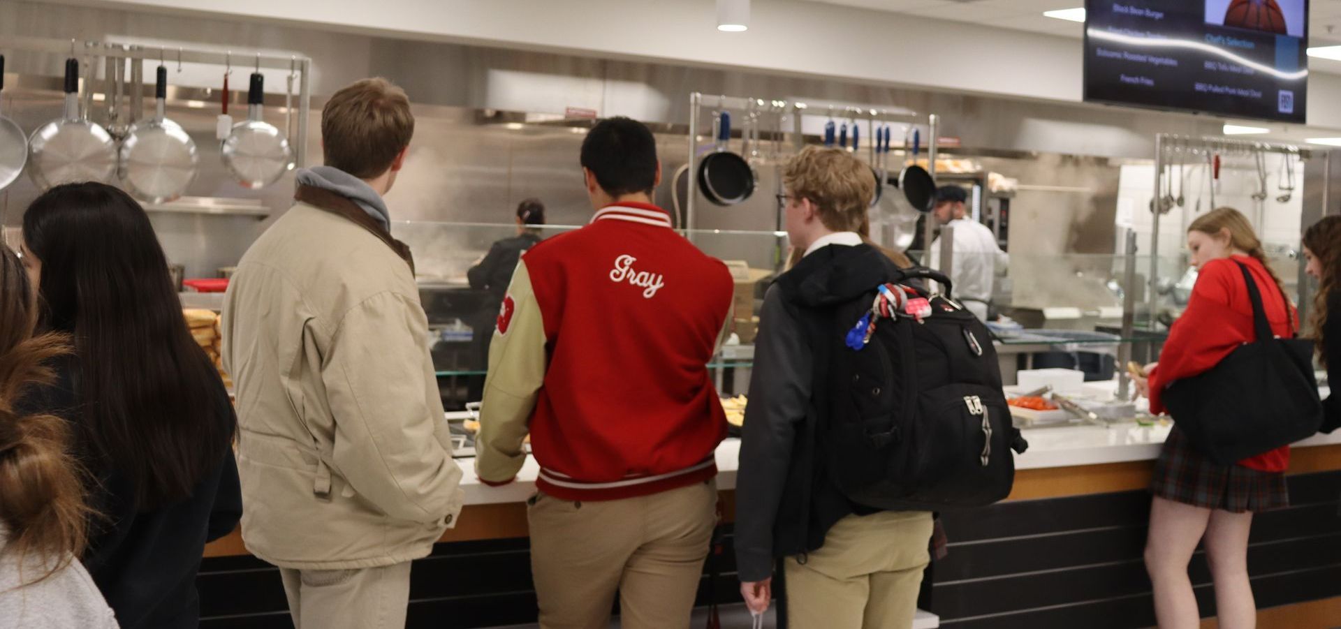 People stand in a dining hall food line, looking at food being prepared by staff behind the counter.
