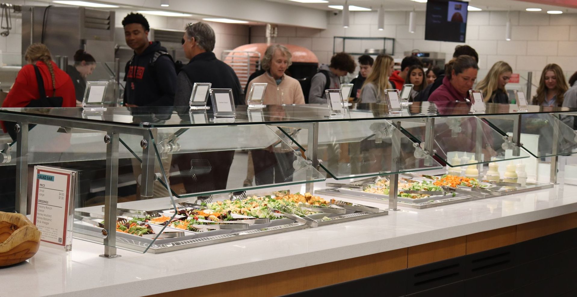 People at a dining hall food bar. Salad bar in the foreground; customers line up to get food.