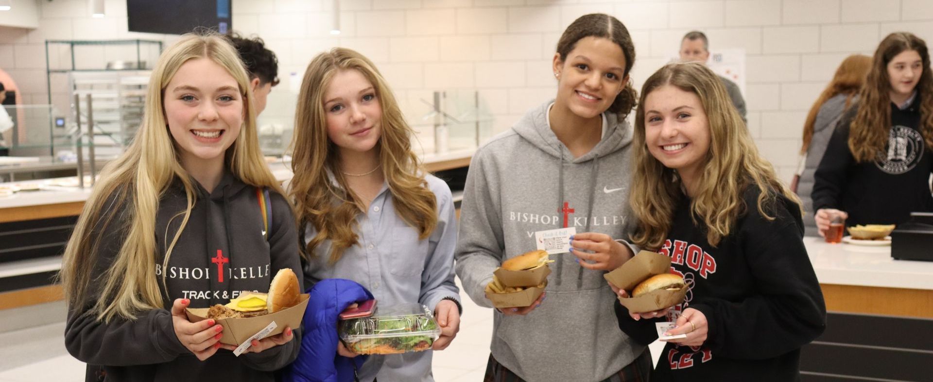 Four smiling female students holding food and food containers in a school dining hall.