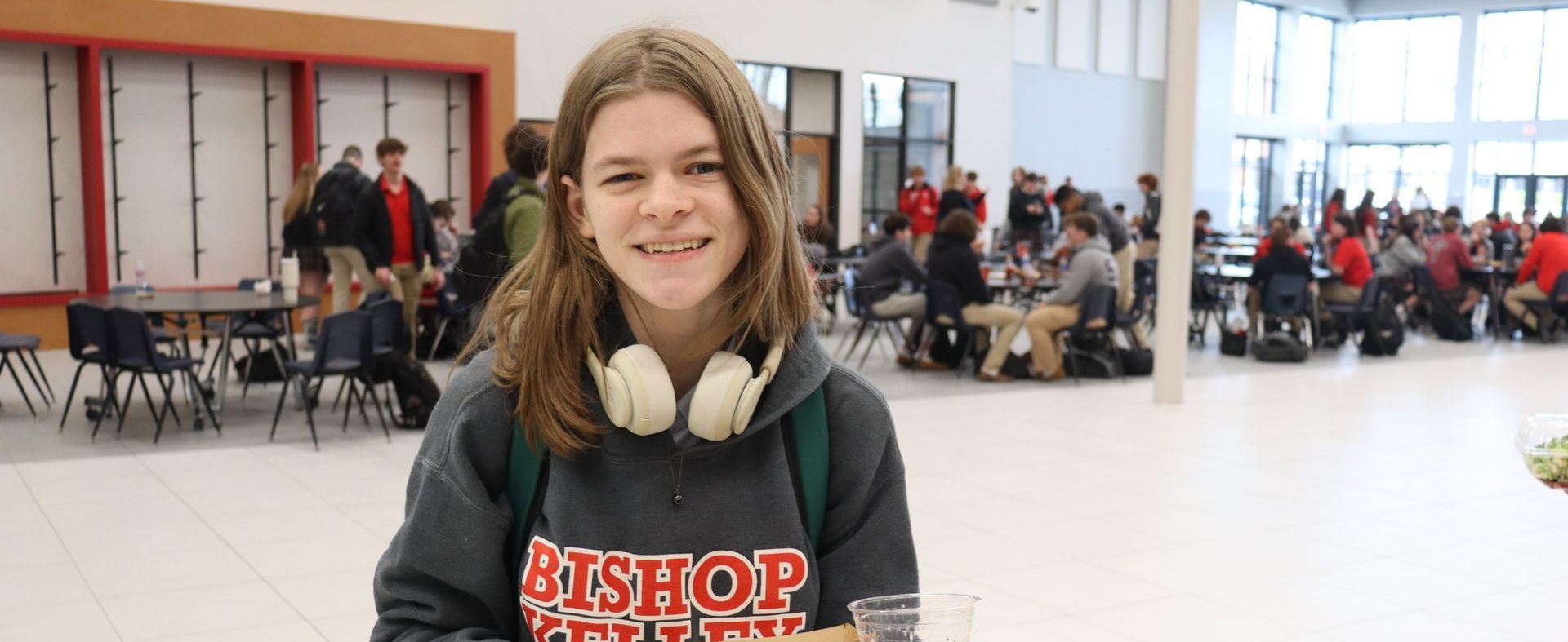 Teen with headphones smiles, posing in a school dining hall. Students sit at tables in the background.