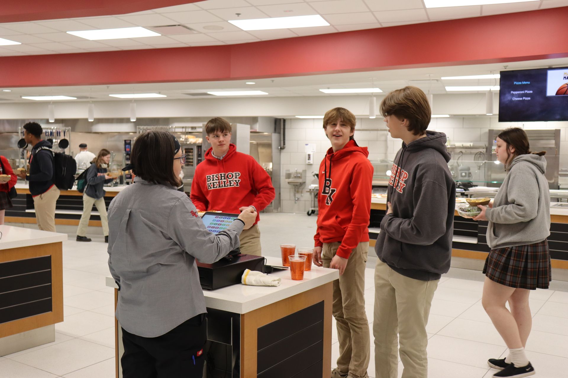 Students in a school dining hall; one pays a cashier. Others stand nearby and wait.