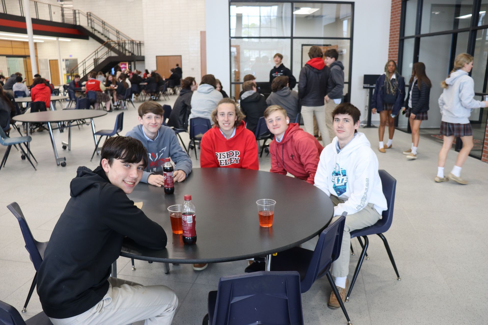 Five students sit at a table in a school dining hall, smiling. Other students and tables are in the background.