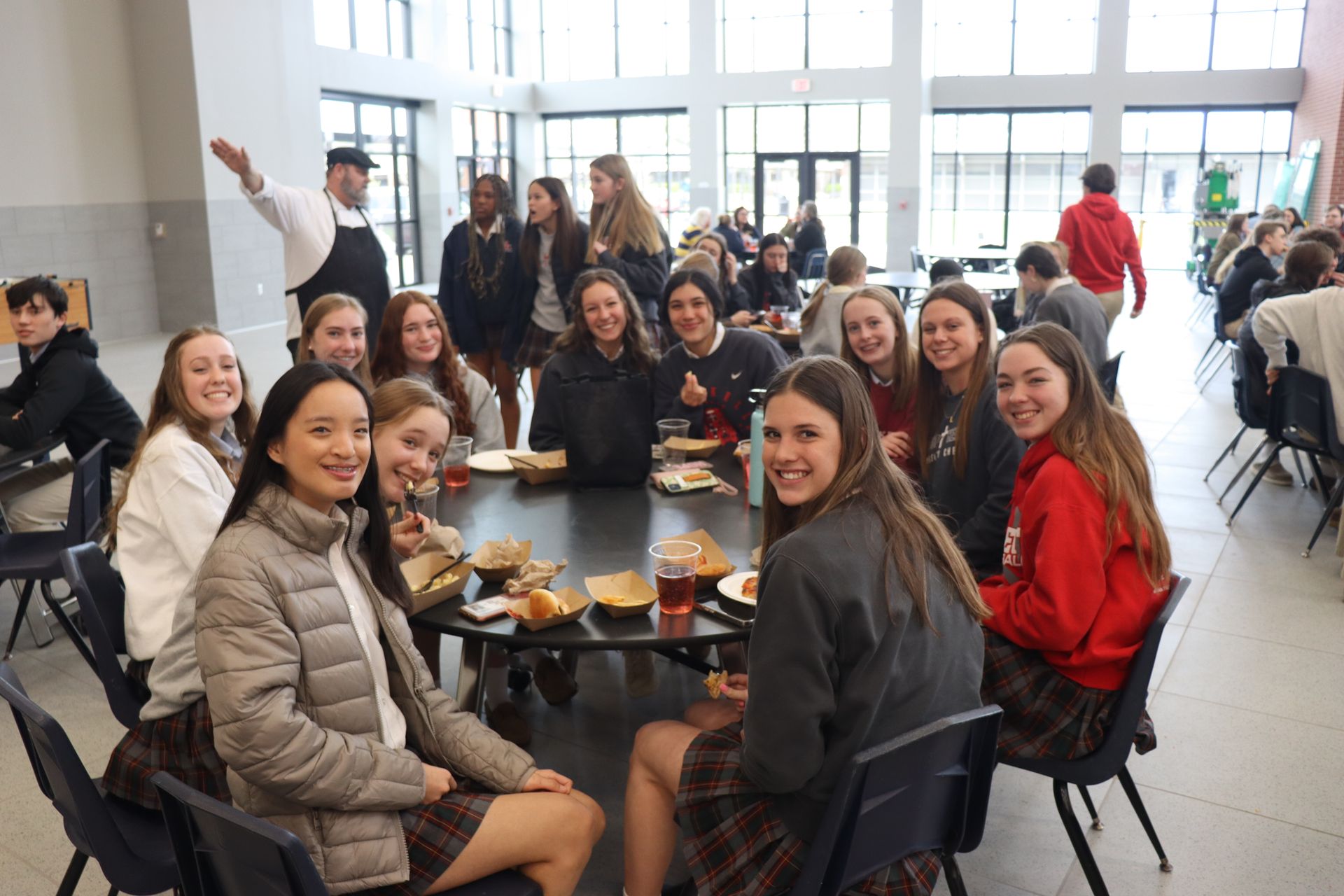 Teenagers eating at a table in a dining hall, some looking at the camera. A cook gestures in the background.