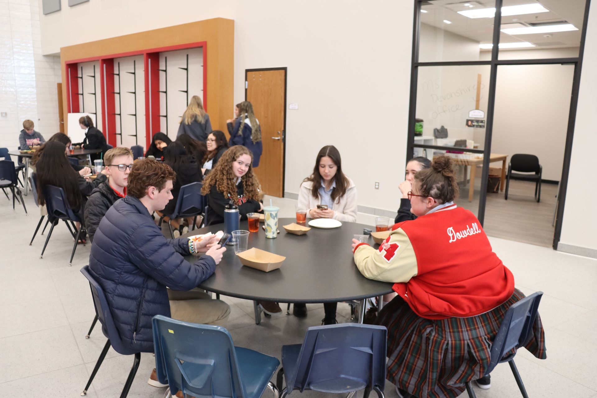 Students sit and talk at a round table. A woman wears a red jacket; a window is in the background.