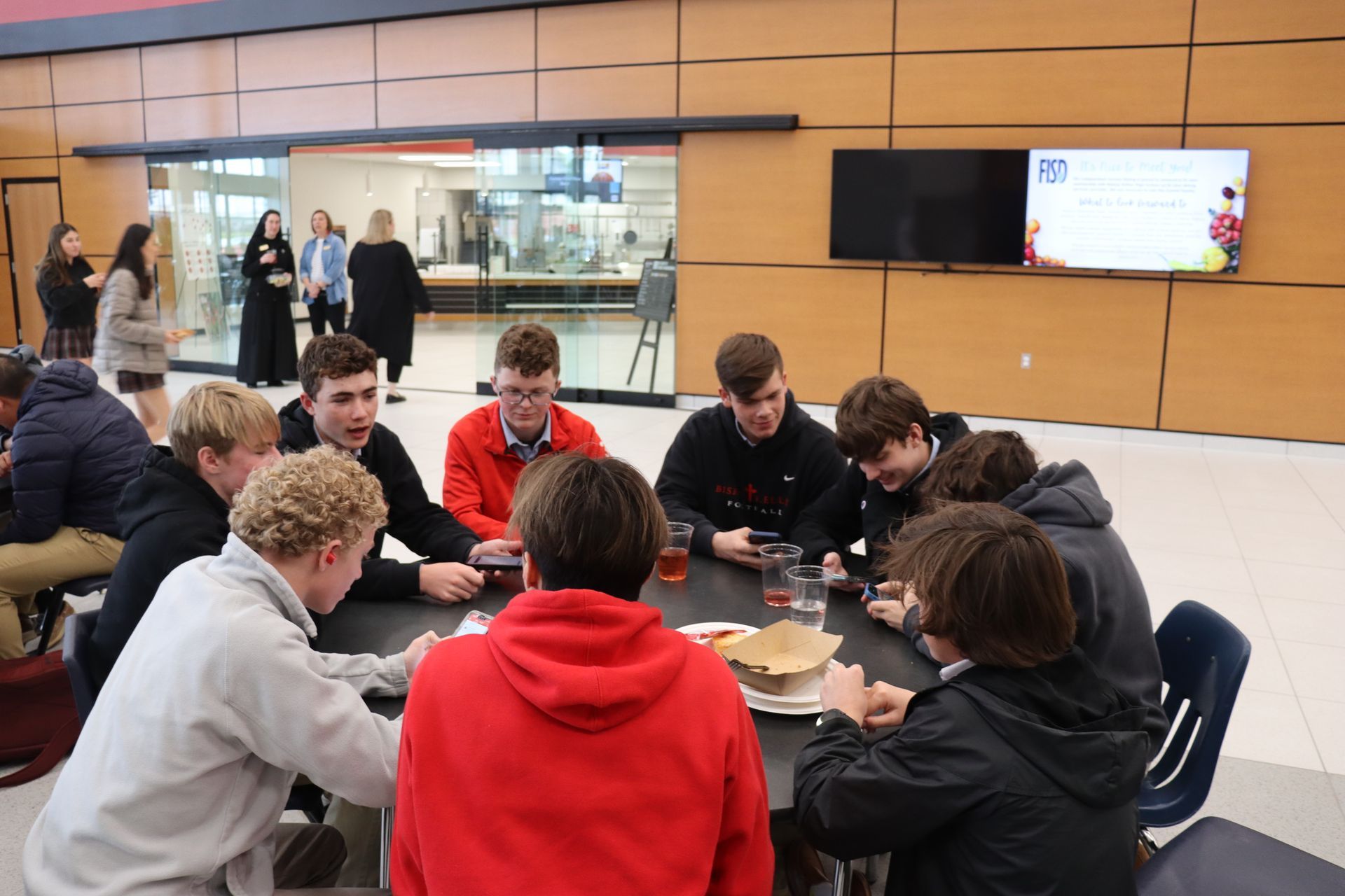 Group of students at a table in a dining hall, looking at their phones and food.