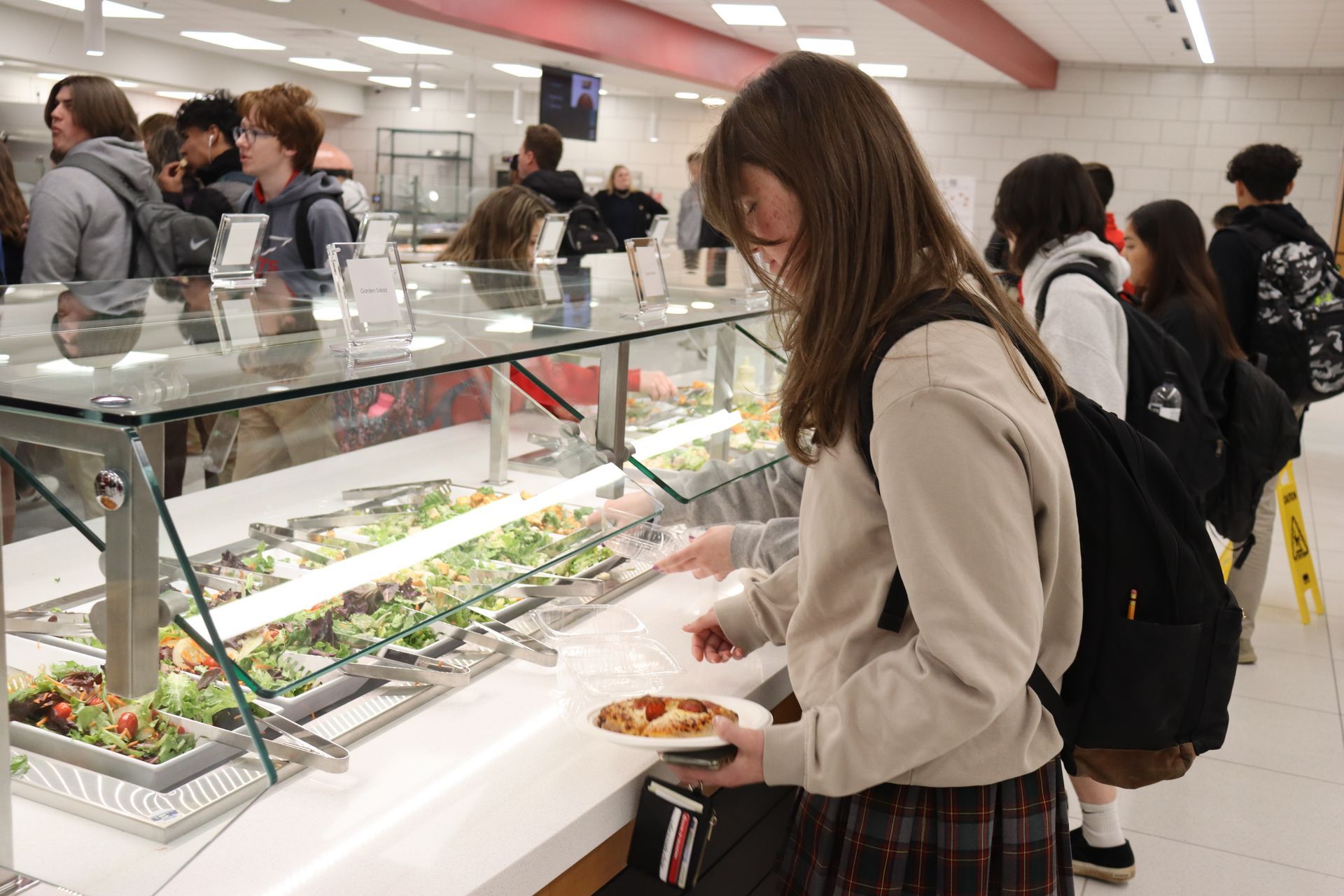 Students line up at a school dining hall food bar, selecting food. Salad bar visible.