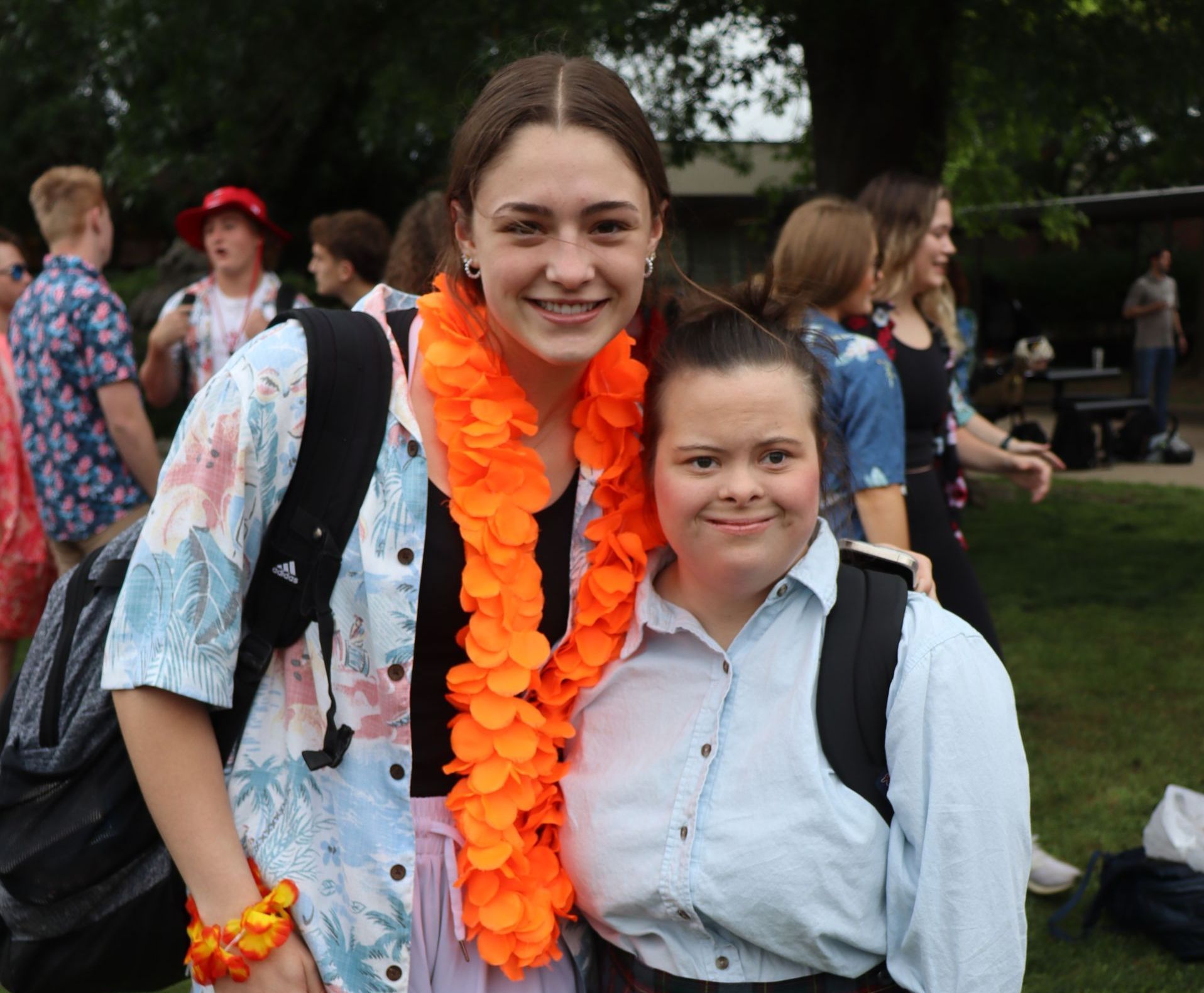 Two smiling women with backpacks pose outdoors. One wears an orange lei and floral shirt. The other has a button-up shirt.