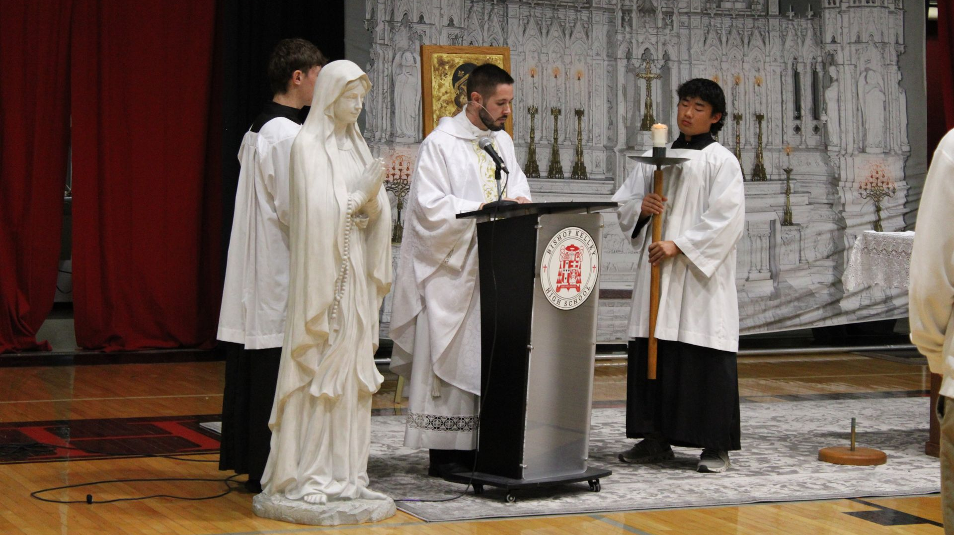 All-School mass. A priest in red and white robes, students, altar, and red backdrop.