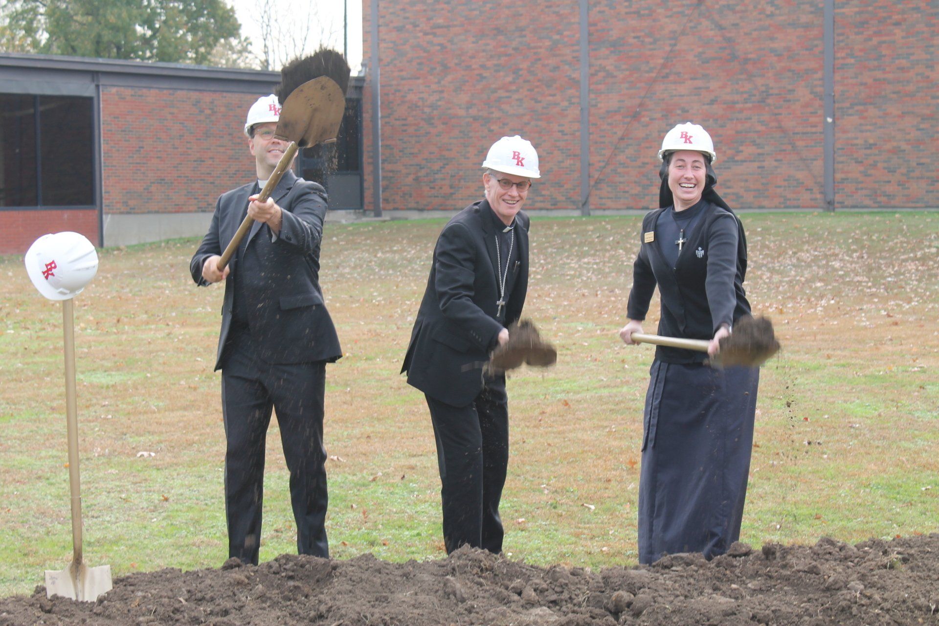 Three people in hard hats and dark clothing shovel soil at a construction site.