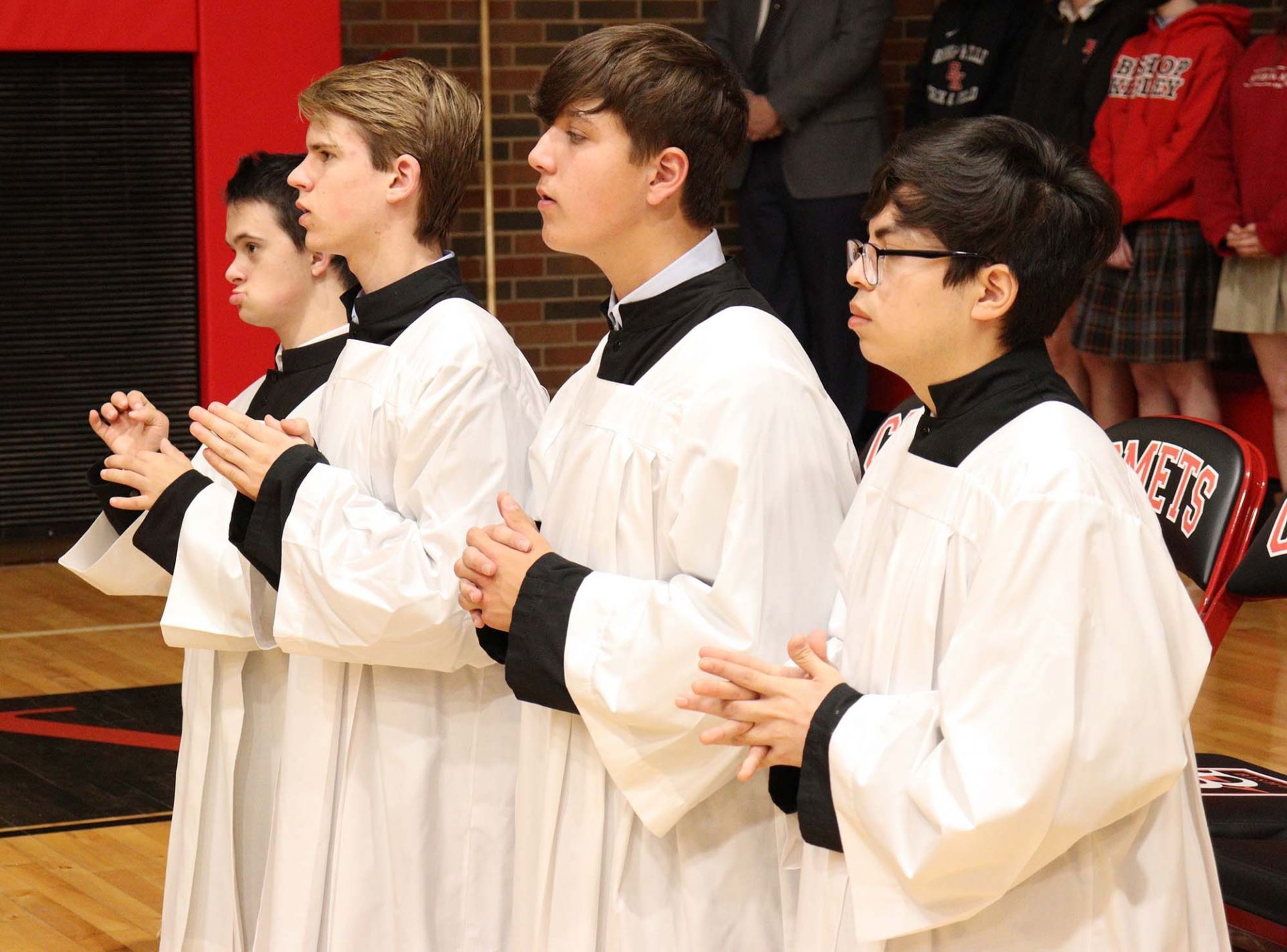 Three people in white robes standing in a gym, hands clasped in front of them.