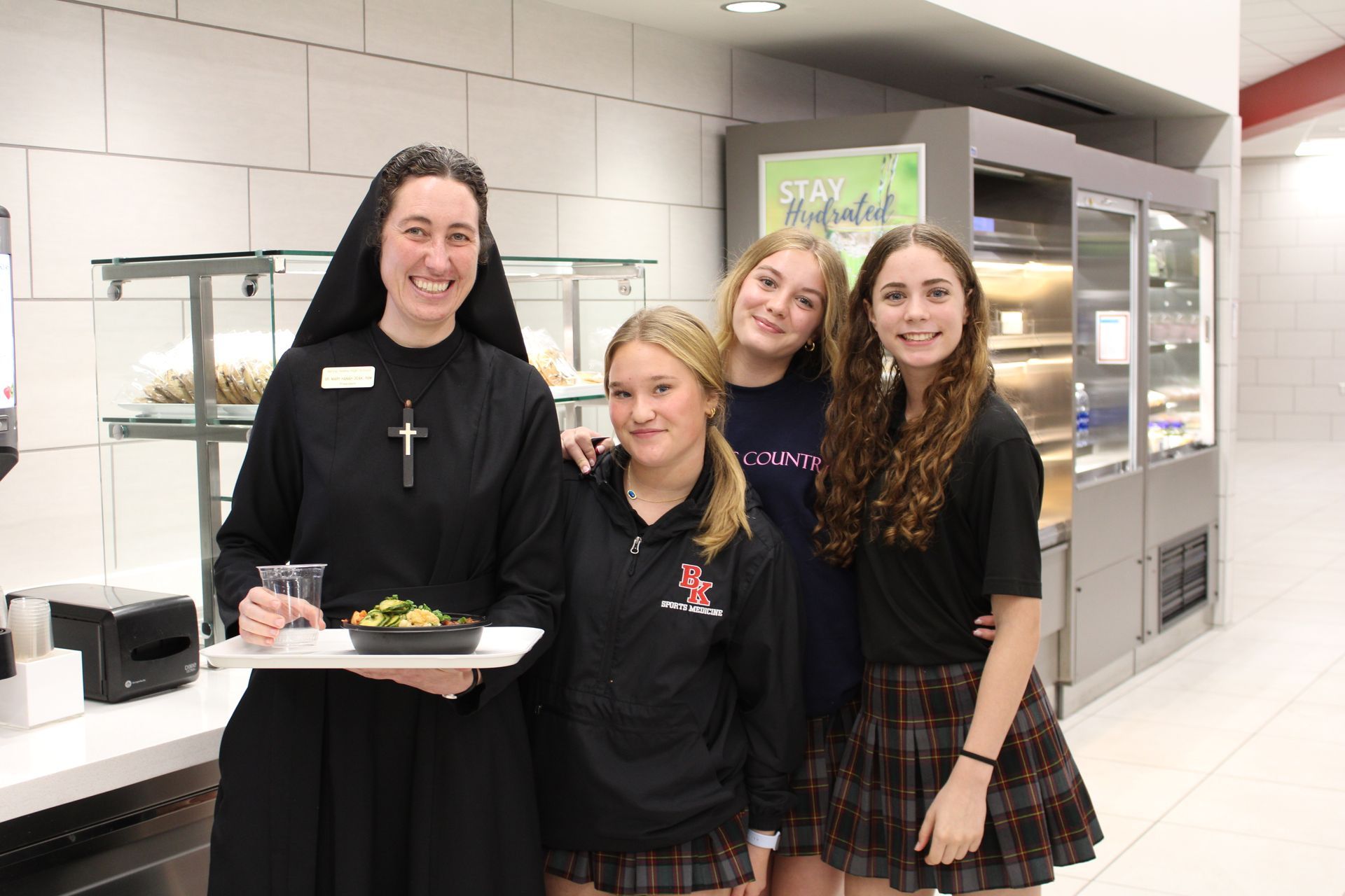 Religious Sister of Mercy and three students smiling in a school dining hall. The nun holds a tray of food.
