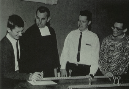 Four men leaning over a table, possibly doing a science experiment. They are smiling and looking at something.