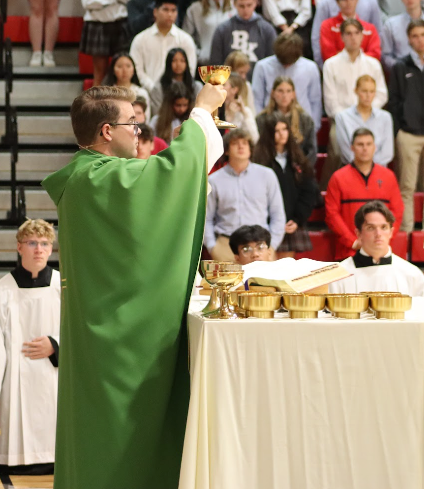 Priest holding chalice during service, surrounded by students in a school setting.