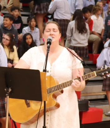 Woman in white dress sings, plays acoustic guitar, indoors. Audience watches in bleachers.