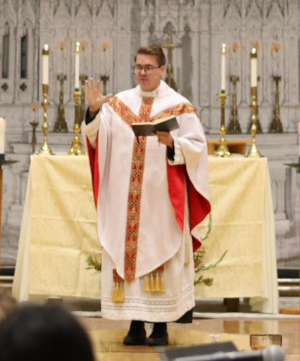 Clergyman in white and red vestments, reading from book, raising hand at altar in a church.