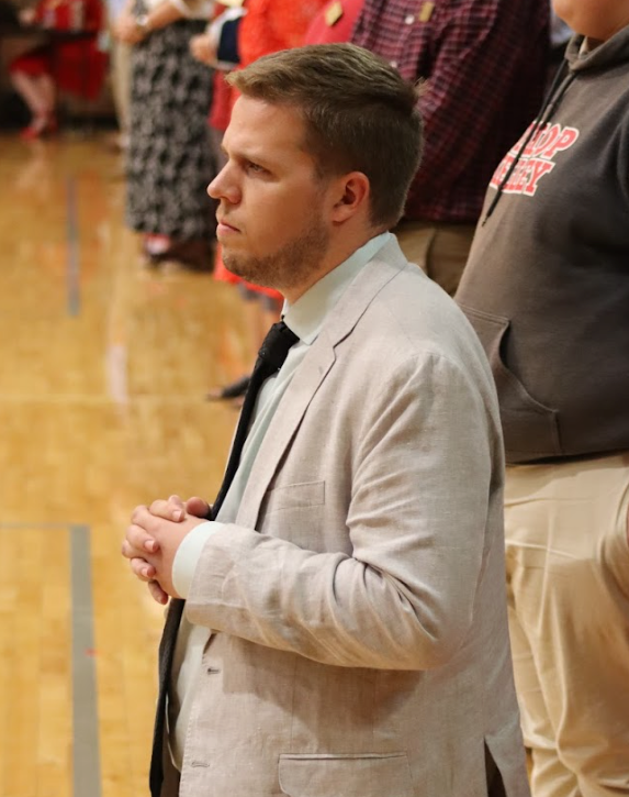 Man in light-colored suit kneels indoors, hands clasped. Others in the background.