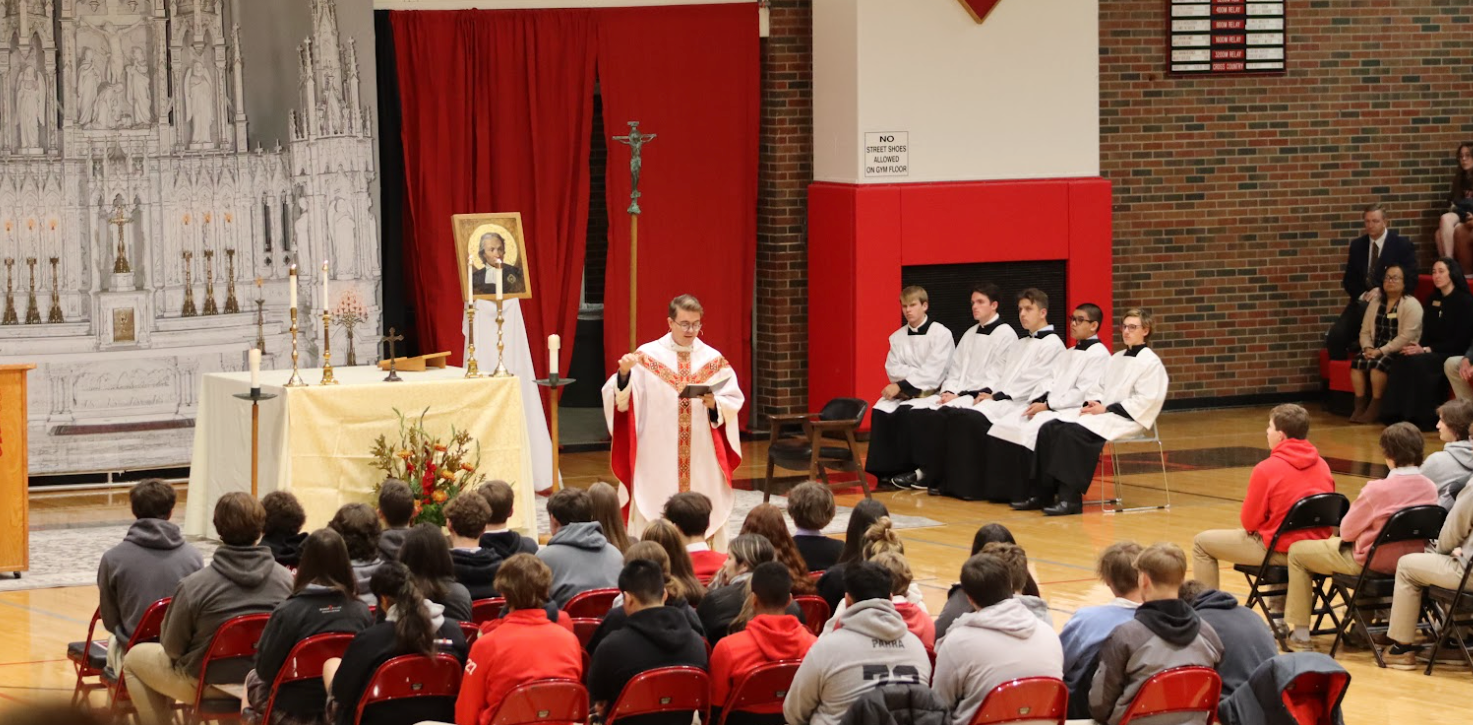All-School mass. A priest in red and white robes, students, altar, and red backdrop.