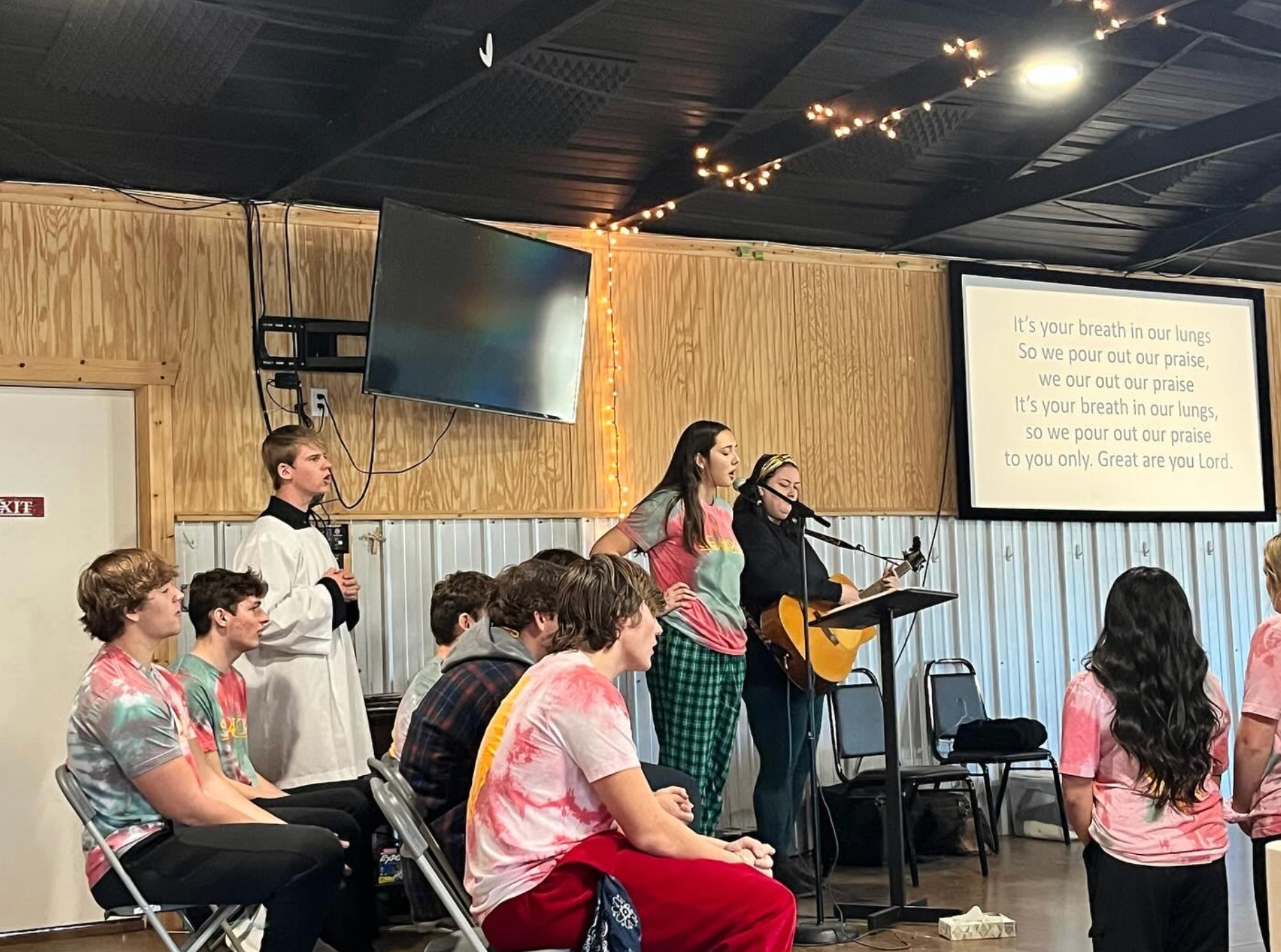 Group of teens singing and watching a performance with lyrics on a screen. Indoors with wood paneling.