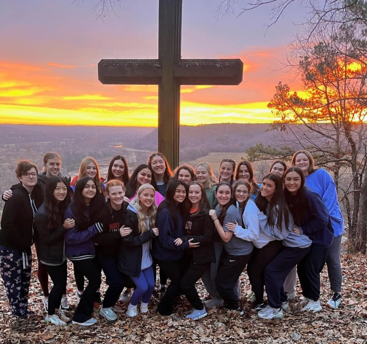 Group of young adults posing under a wooden cross against a colorful sunset.