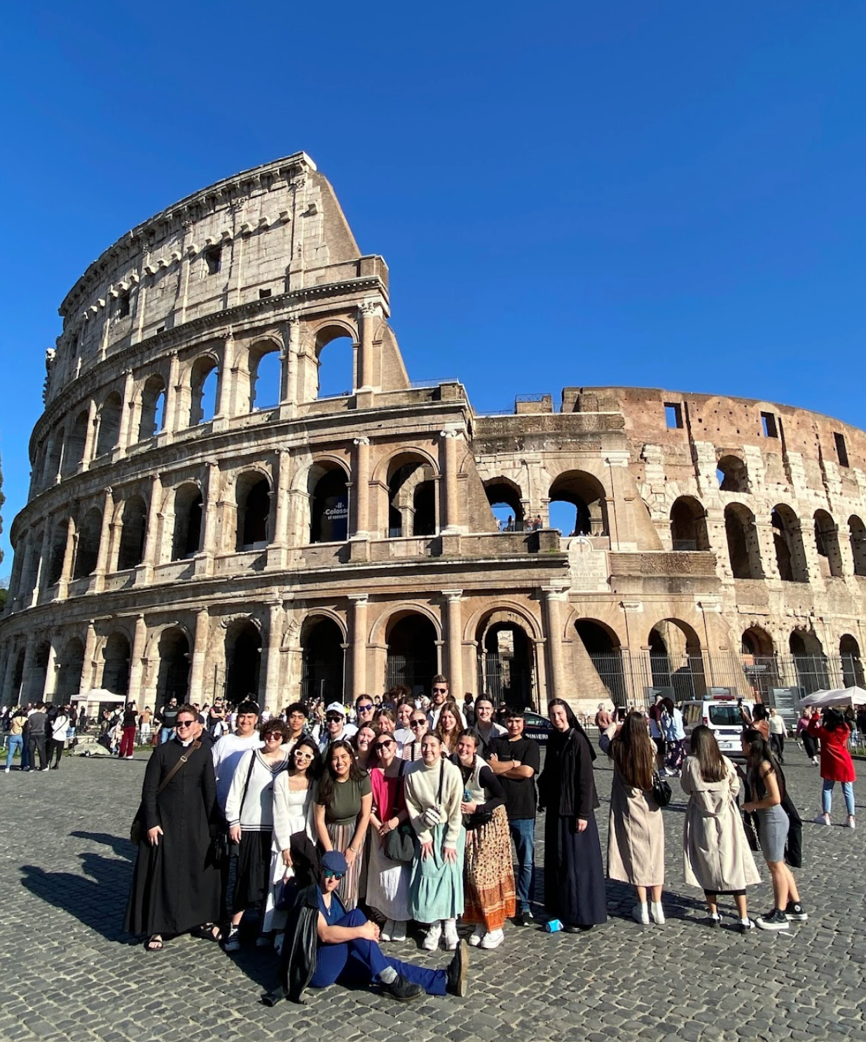 Group of people posing in front of the Colosseum in Rome under a bright blue sky.