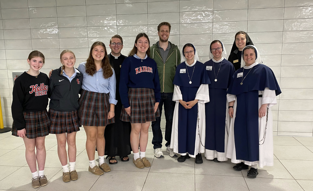 Group of people, including individuals in plaid skirts and nuns, posing in front of a textured white wall.