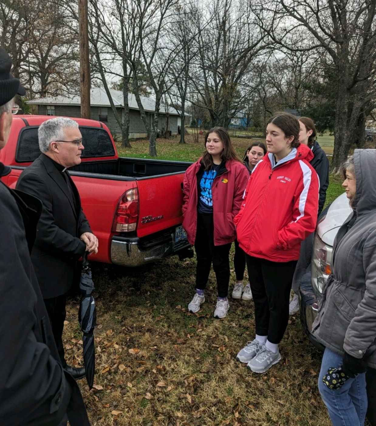 Man in suit talking to a group of teens in front of a red truck and a building on a cloudy day.