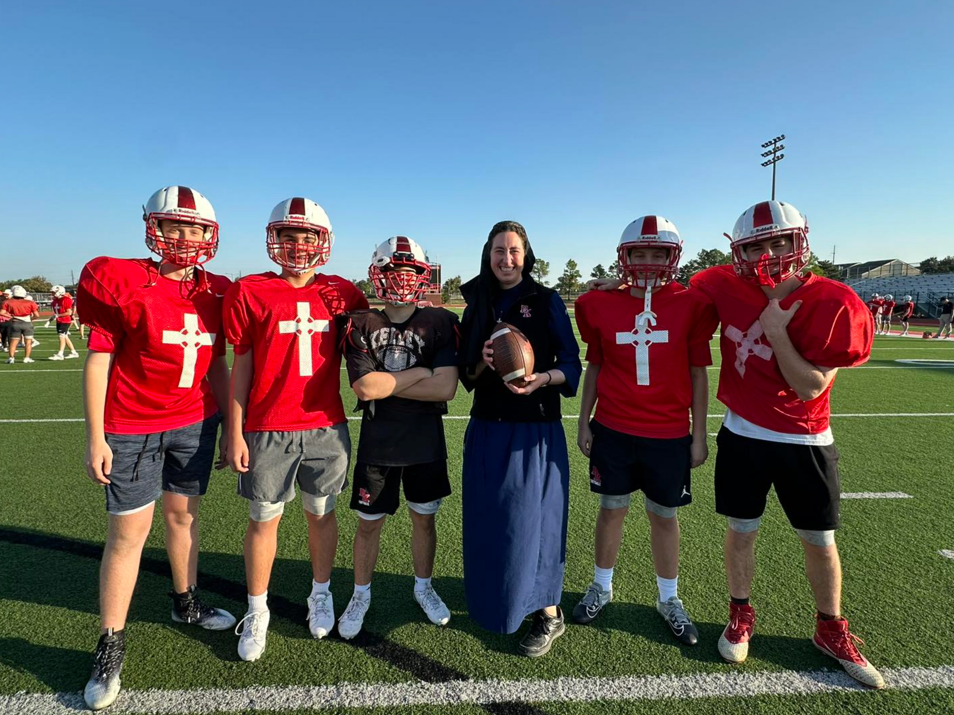 A Religious Sister of Mercy in a habit holding a football poses with five football players on a field.