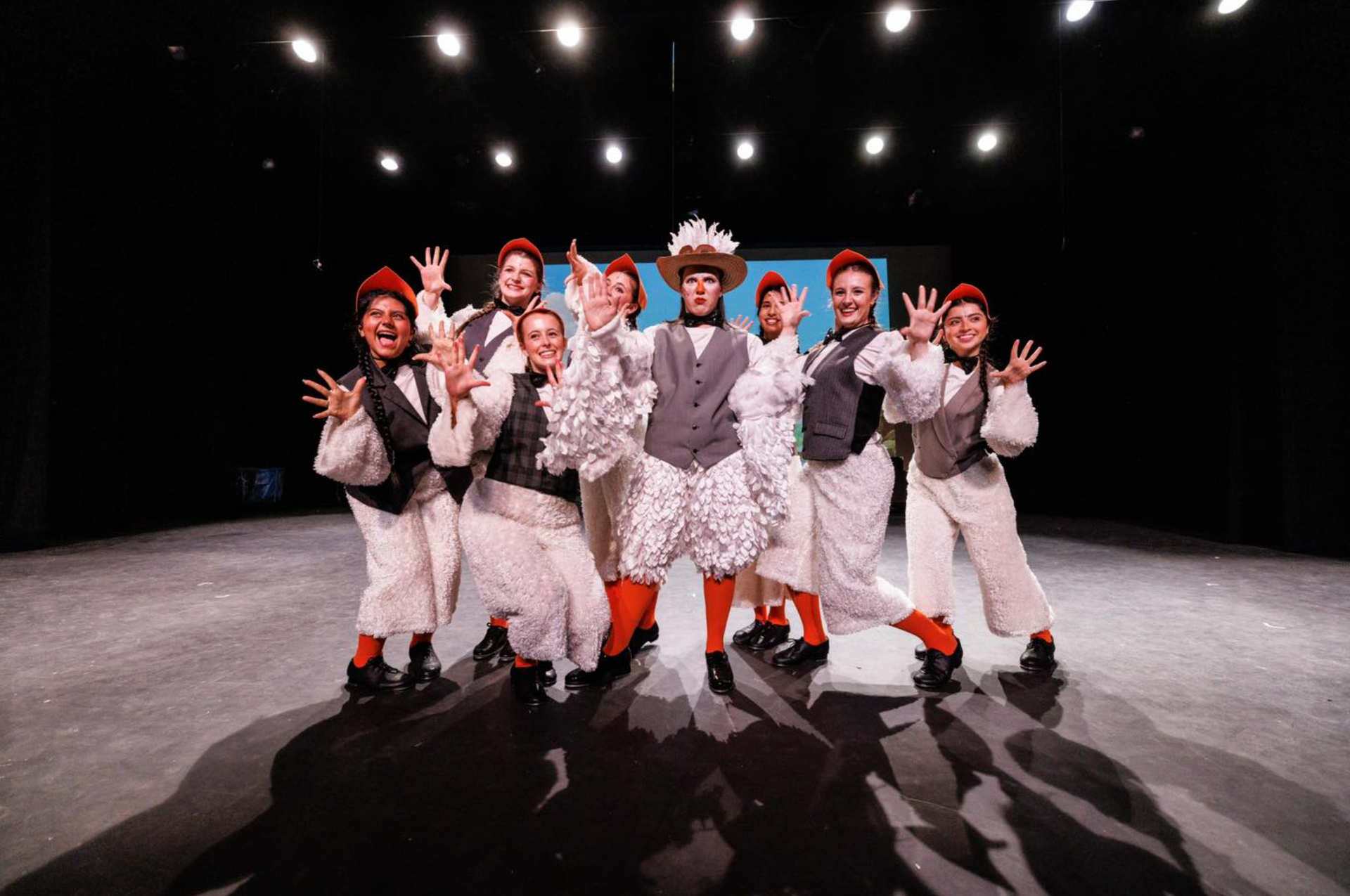 A group of costumed actors on a stage with outstretched hands, smiling.