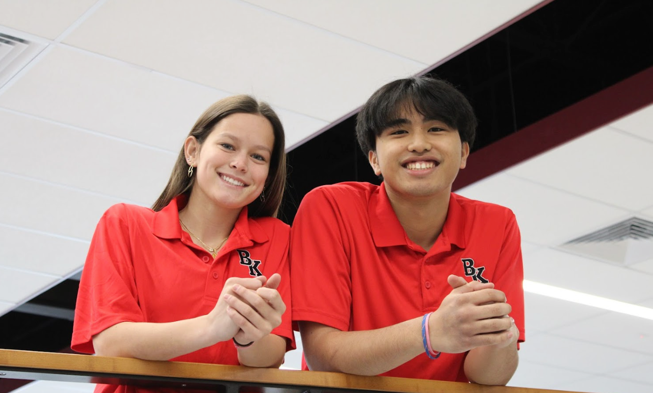 Two smiling people in red shirts lean on a railing, indoors.
