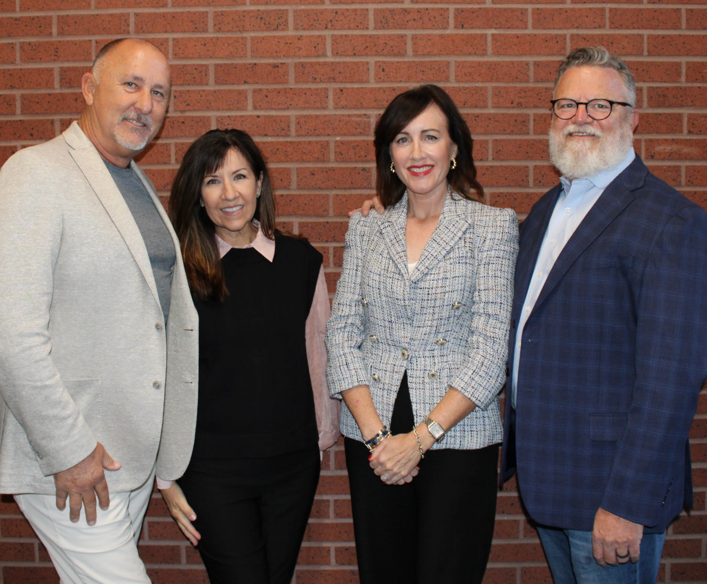 Four people standing in front of a brick wall, smiling. Two women and two men wearing business attire.