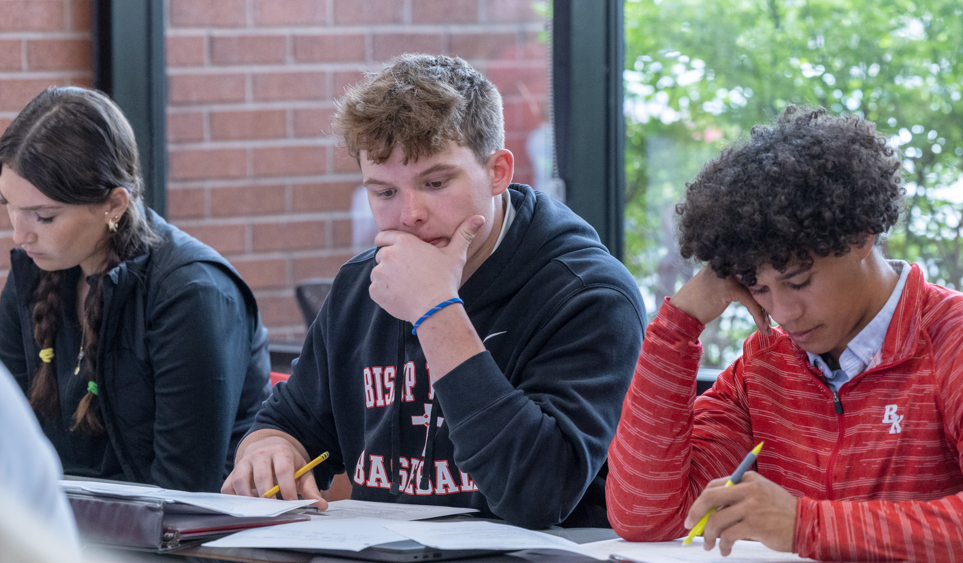 Students in a classroom, looking at papers. One in a black sweatshirt touches chin, others write. Brick wall background.