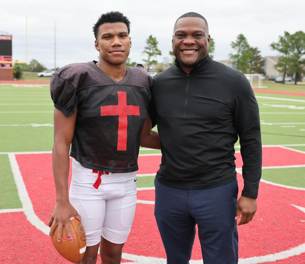 Football player in a jersey with a red cross stands with a man in a black jacket on a football field.