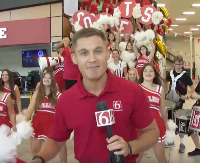 Reporter in red shirt, holding microphone, in front of a cheerleading squad with pom-poms, letters, and drums.
