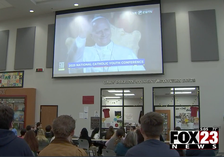 Students watch a screen showing the Pope at a conference. The setting is a large room with tables.