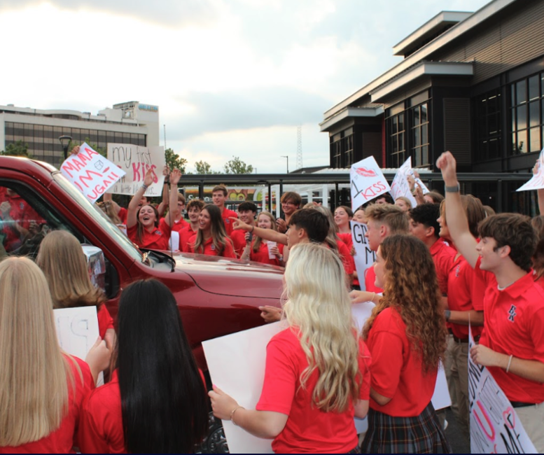 Students in red shirts cheer around a red truck, holding signs outside a building.