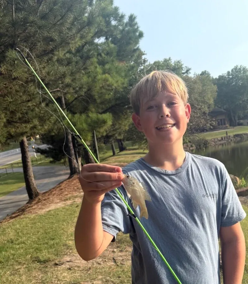 Smiling child holding a fish on a fishing line outdoors near trees and a road
