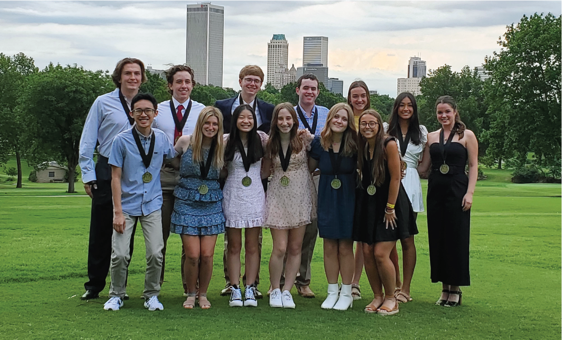 Group of teens in formal wear with medals pose on a grassy field; city skyline in background.