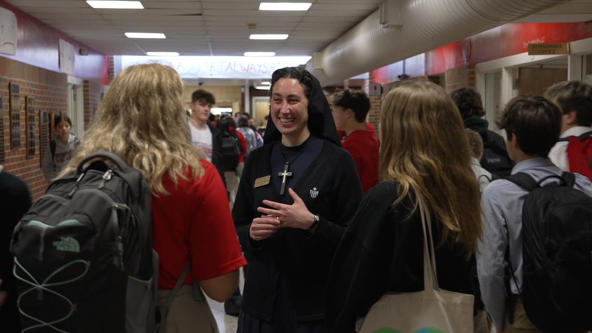 Students chatting in a school hallway, with a woman in a black hoodie speaking at center.