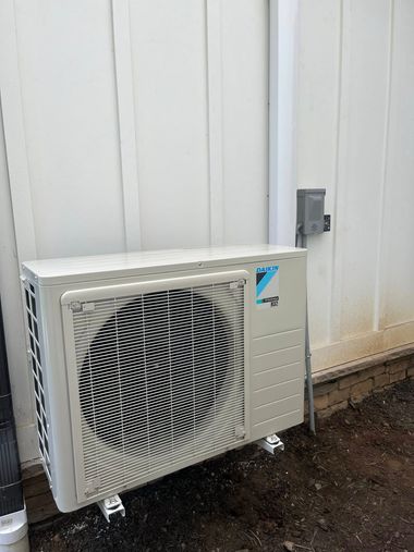 A beige Daikin outdoor HVAC unit mounted on a light wall next to an electrical disconnect box and a vertical white conduit.