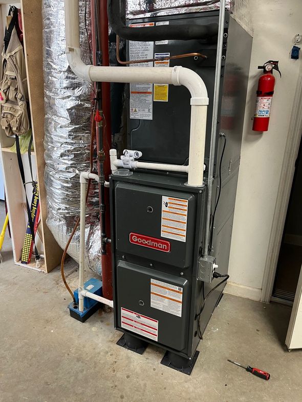 A grey residential furnace stands in a basement next to white PVC exhaust pipes, a metal support pole, and wiring.