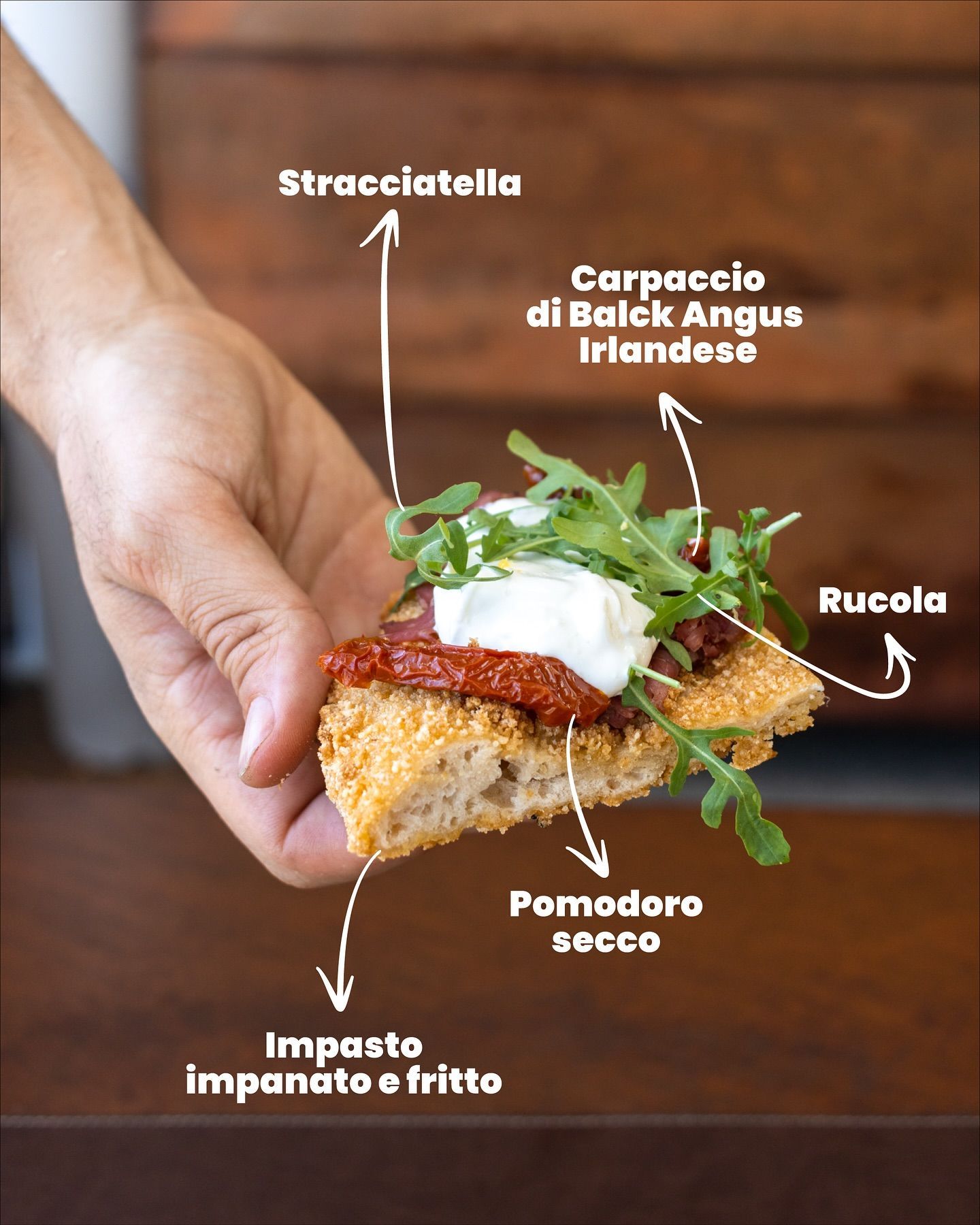 Mano che tiene una fetta di pane con stracciatella, rucola, pomodori secchi e carpaccio.
